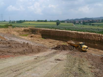 Abbau von Löss und Lösslehm Blick von oben in eine große Lehmgrube. An der Abbauwand sind dunkel- und hellbraune Schichten erkennbar. In der Grube befindet sich ein gelber Bagger. Es schließt sich grüne Landschaft mit Wiesen und einzelnen Bäumen an die Grube an.