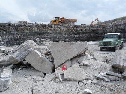 Lockerungssprengung im Böhringen-Sulfat Blick auf Sohle und hintere Wände eines Steinbruches. Im Vordergrund liegen helle Gesteinsbrocken über- und aufeinander. Rechts steht ein Jeep. Im Hintergrund, auf der nächsten Sohle, sind ein Bagger sowie ein Ladefahrzeug erkennbar.