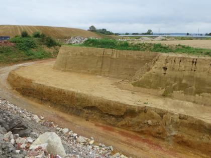 Abbau von quartärem Lösslehm über Deckenschottern und Schichten der Molasse. Blick in eine Tongrube. Über einem halbhohen Socke aus rötlich braunen Erdschichten erhebt sich eine hohe, gelblich braune Abbauwand. Im Hintergrund sind Abraumhügel und Gesteinstrümmer erkennbar.