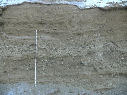 Lagen aus Albschutt mit unterschiedlichen Korngrößen Blick auf eine graubraune Abbauwand. Waagrecht in der Mitte eine Lage Schotter. Links lehnt ein Maßstab an der Wand. Die Kuppe der Wand ist mit Schnee bedeckt.