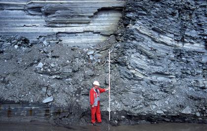 Abbauwand mit dunklen Ton- und grauen Mergelsteinen im Steinbruch Dormettingen Das Bild zeigt die Abbauwand eines Steinbruches. Das blaugraue Gestein ist plattig und scherbig. In der Bildmitte hält ein Arbeiter mit Schutzweste und Helm einen Maßstab hoch. Vor der Wand steht Wasser.