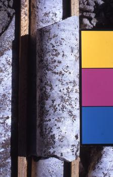 Karbonatit vom Badberg (Kaiserstuhl) Das Foto zeigt einen runden, an den Enden abgeschrägten Steinkörper aus einer Bohrung. Der Stein ist weißlich grau mit braunen Flecken. Der Stein liegt auf anderen, in Kisten gelagerten Bohrkernen. Rechts ist eine farbige Markierung angebracht.