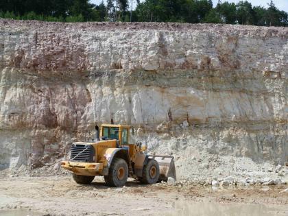 Stubensandsteingrube bei Kernen im Remstal Blick auf eine Steinbruchwand mit drei aufeinanderliegenden, durch grünliche Streifen getrennte Schichten. Die Farbe des Gesteins ist ein weißliches Grau, links sind jedoch auch violette Farbtöne zu sehen. Im Vordergrund ein gelber Radlader.