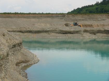 Rheingletscher-Niederterrassenschotter im Trocken- und Nassabbau Blick auf die waagrecht verlaufende Wand einer Kiesgrube mit Schaufellader und vorne anstehendem See. Links schiebt sich eine weitere Abbauwand ins Bild.