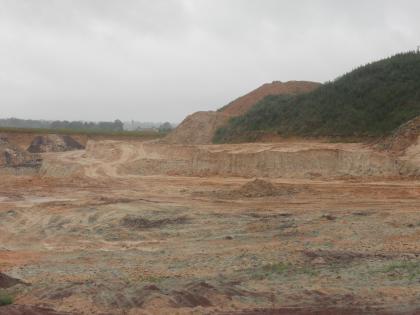Abbaukante der plattig aufgewitterten, mürben Sandsteine. Blick auf eine im Mittelgrund sich erhebende Abbaukante in einer Sandgrube. Im Vordergrund die abgeflachte Grube. Im Hintergrund rechts steigen bewachsene Hügel und Abraumhalden auf.
