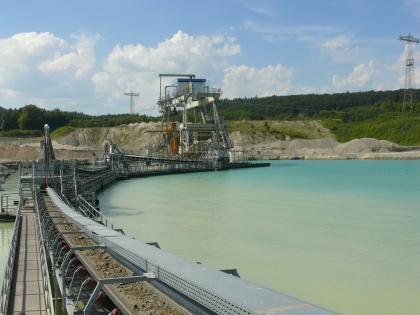 Nassabbau mittels Schwimmbagger Das Foto zeigt in der Bildmitte einen Schwimmbagger auf einem See. Mehrere Förderbänder führen in einem Bogen links vom Bagger auf den Betrachter zu. Im Hintergrund Abraumhalden und Wald.