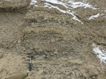 Sandige, fein- bis grobkiesige, schwach steinige Schotter Nahaufnahme von kleineren und größeren Kieselsteinen, in graubraunes Bodenmaterial verbacken. Oben und rechts liegt etwas Schnee.