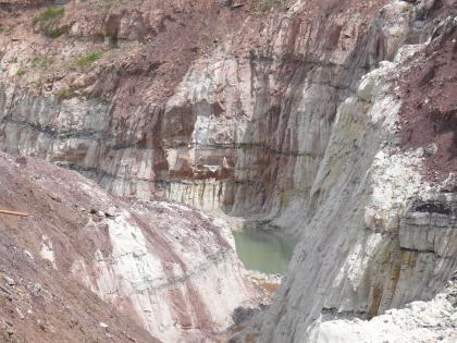 Sandsteine im Steinbruch Berglen-Hößlinswart Das Bild zeigt im Hintergrund und rechts leicht geneigte, weißlich graue bis violette Steinbruchwände. Am Fuß ist eine kleine Wasserfläche erkennbar. Links vorne schieben sich Abraumhügel ins Bild.