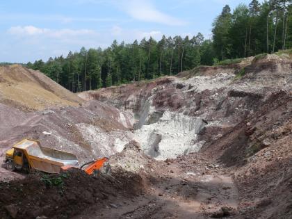 Abbau des Stubensandsteins im Steinbruch Berglen-Hößlinswart Von erhöhtem Standort aus überblickt man rechts Steinbruchwände und links Abraumhalden. Links unten wartet ein gelber Kipplaster auf Beladung. Im Hintergrund bildet ein nach rechts ansteigender Wald den Abschluss des Bildes.