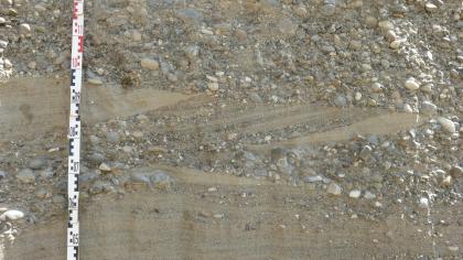 Nahaufnahme mehrerer Schichten von feinem, hellbraunem Sand im unteren Drittel und grobem Kies im oberen Drittel. Die Schicht in der Mitte zeigt sowohl Kies als auch Sand und verläuft schräg zur ansonsten waagrechten Linie.