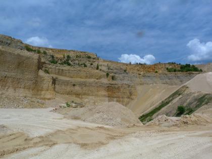 Auskeilende, dunkelgraue Lacunosamergel (joL) Der Betrachter befindet sich in einem Steinbruch aus hellbeigem Gestein, welches auf mehreren Sohlen abgebaut wird. Die obere Bildhälfte wird von intensiv blauem Himmel eingenommen.