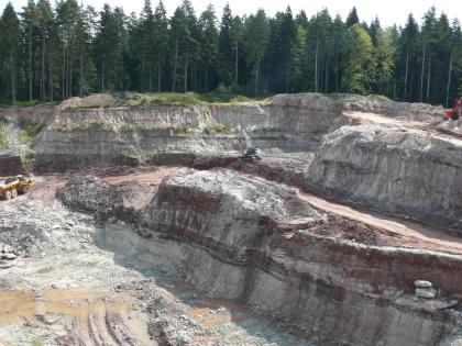 Abbauwand einer Mürbsandsteingrube bei Gschwend Blick von erhöhtem Standort auf mehrere hintereinanderliegende, violett-graue Steinbruchwände. Die Brüche sind durch Fahrstraßen getrennt. Im Hintergrund stehen hohe Nadelbäume an.