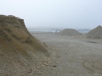 Kiesgewinnung im Trocken- und Nassabbau Das Bild zeigt größere Kiesberge links vorne und rechts hinten. Die Berge stehen auf ebenem, trockenem Boden. Im dunstigen Hintergrund sind ein See sowie Förderbänder zu erkennen.