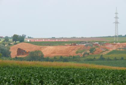 Abbau von quartärem Lösslehm und aufgearbeitete Tonsteine der Grabfeld-Formation. Inmitten einer grünen Nutzlandschaft befindet sich in der Bildmitte eine Lehm- und Tongrube. Das anstehende Gestein ist rötlich-braun.