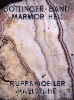 Musterplatte der hellen Varietät von Böttinger Marmor Musterplatte von hellem Gestein mit der Aufschritt „Böttinger Bandmarmor Hell“. Zwischen dem hellen Gestein verlaufen senkrechte, leicht nach rechts geneigte dunklere Streifen.