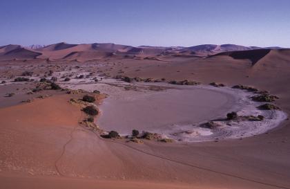 ausgetrockneter, verflochtener Fluss (braided river) in der namibischen Wüste Das Bild zeigt eine afrikanische Wüste mit Dünen im Hintergrund und einem gewundenen, beinahe ausgetrockneten Fluss im Vordergrund. Der Fluss endet in einem runden, flachen Becken.