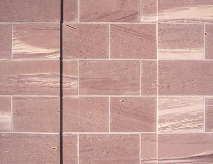 Nahaufnahme einer Hausfassade mit rötlichen, durch helle Fugen getrennten Mauersteinen. Links und vor allem rechts sind die rötlichen Steine mit hellen Streifen versehen; teils sind auch schräg verlaufenden dunkle Streifen erkennbar.