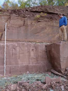 Rote, bankige bis dickbankige Feinsandsteine im Steinbruch Tiefenbronn bei Mühlhausen Zu sehen ist hier eine rötlich graue, glatte hohe Steinbruchwand. Rechts ragt ein großer Block heraus, auf dem ein Mann steht. Vor der Wand liegen Bruchscherben. Links lehnt eine Messlatte an der Wand.