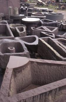 Brunnen und Pflanztröge für den Landschafts- und Gartenbau, hergestellt aus Plattensandstein Das Foto zeigt mehrere auf dem Boden abgestellte, aus rötlich grauem Stein gefertigte Brunnen und Pflanztröge.