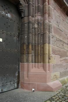 Neuer Sockelstein am Breisacher Münster Seitenansicht eines Toreingangs aus rötlichem, am Tor schwärzlichem Mauerwerk. Auffallend ist ein neu eingesetztes Sockelstück aus rosafarbenem Gestein.