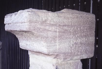 Balkenwiderlager aus grobkörnigem, schräg geschichtetem Degerfelder Sandstein Blick auf einen größeren, bearbeiteten Steinblock. Der Stein hat eine rötlich graue Farbe und liegt auf einem zweiten Steinblock auf. Links hat der Block eine runde, nach unten zeigende Ausbuchtung.