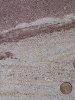 Typisch grober Buntsandstein aus Degerfelden Nahaufnahme eines Gesteins, welches in den unteren zwei Dritteln des Bildes hellbeige ist und eine leichte Schichtung aufweist. Im oberen Drittel ist das Gestein dunkelrot. Rechts unten befindet sich eine Münze als Maßstab.