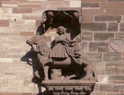 Reiterstatue am Basler Münster Das Foto zeigt eine aus Stein gehauene Reiterstatue mit Sockel und Überdachung. Statue sowie das dahinter sichtbare Mauerwerk eines Gebäudes bestehen aus rötlich grauem Gestein.