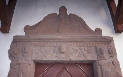 Renaissanceportal aus Schopfheimer Plattensandstein an der evangelischen Kirche in Schopfheim Blick auf die aus Stein gefertigte Einfassung einer Tür. Das Gestein ist rötlich grau und reich verziert; auf dem Sims befindet sich zusätzlich ein bogenförmiger Aufsatz.