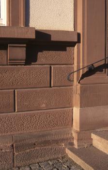Sockel- und Mauersteine aus Sandstein am Schopfheimer Rathaus Blick auf den Eingangsbereich eines Gebäudes. Sockel (links), Fensterbrüstung (links oben) und Treppe (rechts) bestehen aus rötlich grauen, teils porigen Steinen. Die Fenstereinfassung sowie die Treppenwand sind aus glatterem Gestein gefertigt.
