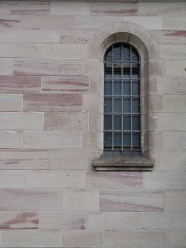Fensterpartie der Herz-Jesu-Kirche in Gernsbach-Obertsrot Das Foto zeigt ein schmales vergittertes und oben abgerundetes Fenster in einer rötlich grauen Steinmauer.