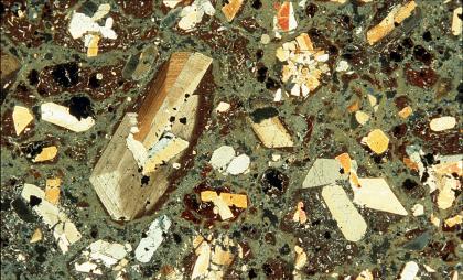 Dünnschliffbild eines Phonoliths Das Dünnschliffbild zeigt gelblich-weiße bis braune längliche Kristalle, die sich in einer teils rostbraunen, teils graugrüner Grundmasse befinden.