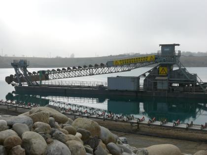 Eimerkettenbagger in einem Baggersee Das Foto zeigt den Transport von Abbaueimern über ein Förderband auf einem schwimmenden Bagger in einem Baggersee. Im Vordergrund sind größere Steine gelagert.