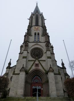 Ansicht des Kirchturms der Kirche St. Konrad in Langenenslingen Blick auf den Eingangsbereich einer Kirche mit sich darüber erhebendem Turm aus grauem Gestein. Der Kirchturm wird von mehreren seitlichen Pfeilern gestützt und dem neugotischen Baustil zugeordnet.