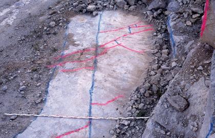 Kluftmuster im Travertin, Steinbruch Gauingen Blick von oben auf eine hellgraue Gesteinsfläche mit blauen und roten Markierungslinien sowie einer Messlatte. Um die Fläche liegt dunkelgrauer Gesteinsschutt, rechts steigen Stufen aufwärts.