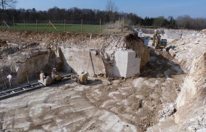 Neuaufschluss im Oberen Lager, Steinbruch Gauingen Blick auf ein flaches, zum Hintergrund hin verzweigtes Steinbruchgelände. In der Bildmitte, an einer vorgelagerten, oben begrünten Insel, finden Brucharbeiten statt.