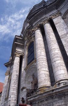 Aufwärts gerichteter Blick auf den oberen Teil einer Kirchenfassade; mit hellgrauen Steinsäulen rechts und links, einer Figurennische und Simsen oben und unten. Der Baustil wird dem Barock zugerechnet.