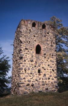 Das Bild zeigt die Front eines viereckigen Turms, welcher auf einer kleinen Erhöhung steht. Der Turm ist aus rötlich bis gelblich grauen Gesteinen gebaut und hat ein großes Fenster und oben noch kleinere Fenster.