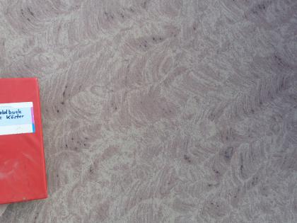 Schichtparallel geschnittener Schilfsandstein Nahaufnahme einer bräunlich grauen Gesteinsoberfläche mit Sägespuren. Links dient ein aufgelegtes Notizbuch als Größenvergleich.