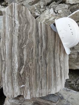 Im Bild ist ein charakteristisch gebänderter Block von Felsensulfat zu sehen. Gipsbruch Deißlingen-Lauffen bei Rottweil Das Foto zeigt einen hellgrau und dunkelgrau gestreiften Gesteinsblock. Die äußeren Kanten sind unregelmäßig gebrochen. Ein weißer Schutzhelm dient als Größenvergleich.