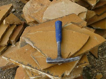 Platten aus Renquishausen-Plattenkalk Auf diesem Foto liegen mehrere orangefarbene, in unterschiedlichen Größen und Formen gebrochene, dünne Gesteinsplatten auf- und nebeneinander. Ein Hammer auf der obersten Platte dient als Größenvergleich.