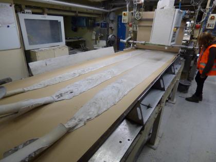 Produktion von Gipskartonplatten Perspektivische Aufnahme einer Maschine mit Auflagefläche. Auf dieser Fläche liegt eine hellbraune, große Platte. Aus drei Schlauchöffnungen links fließt weißlicher, flacher Brei auf die Platte und verteilt sich dort gleichmäßig.