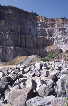 Die Aufnahme zeigt im Vordergrund über- und nebeneinanderliegende hellgraue Steinblöcke. Im Hintergrund ragt die hohe, mehrstufige Wand eines Steinbruches auf.