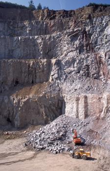Abbau von Flasergneisen, Steinbruch Steinach Blick auf eine sehr hohe, über mehrere Stufen gehende Steinbruchwand aus grauem bis rötlich braunem Gestein. Rechts unten arbeiten Bagger und Radlader vor einer Abraumhalde.