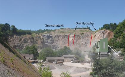Abbau von Gneisanatexiten im Steinbruch Detzeln Blick in einen Steinbruch mit Abraumhalde links vorne, Förderanlagen sowie weiteren Gebäuden und Fahrzeugen rechts und mittig. Im Hintergrund erhebt sich eine hohe Gesteinswand mit farbig hinzugefügten Anmerkungen wie etwa zwei Granitporphyrgängen rechts.