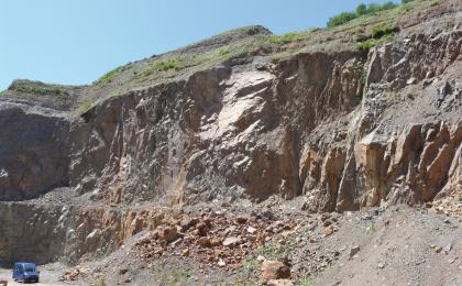 Abbau von Gneisanatexiten im Steinbruch Detzeln Seitenansicht einer Steinbruchwand mit grauem bis rötlichem, mittig waagrecht und außen senkrecht geklüftetem Gestein. Die Kuppe der Bruchwand ist begrünt.
