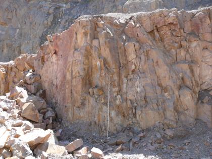 Abbau von Gneisanatexiten mit orthogonalem Kluftsystem im Steinbruch Detzeln Blick auf eine rötlich graue Steinbruchwand, links mit abfallendem, gezahntem Grat sowie abgerutschtem Blockmaterial. Das Gestein ist zudem von feinen Rissen durchzogen. Im Hintergrund ist eine weitere Steinwand erkennbar.