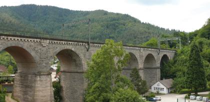 Hornberger Eisenbahnviadukt der Schwarzwaldbahn, mit Forbach-Granit und Raumünzach-Granit verblendet Seitlicher Blick auf eine lange Eisenbahnbrücke aus rötlich grauem Gestein, mit mehreren Pfeilern und Bögen. Im Hintergrund erheben sich bewaldete Berge.