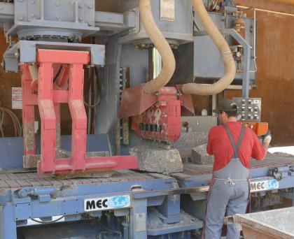 Weiterverarbeitung der Blöcke zu Quadern für Pflaster- und Randsteine mit Hilfe einer hydraulischen Spaltmaschine Das Bild zeigt eine Maschine beim Zerkleinern von Steinblöcken. Erkennbar sind Haltevorrichtungen, ein Laufband und Spaltwerkzeuge. Ein Arbeiter, der dem Fotograf den Rücken zukehrt, bedient die Maschine.