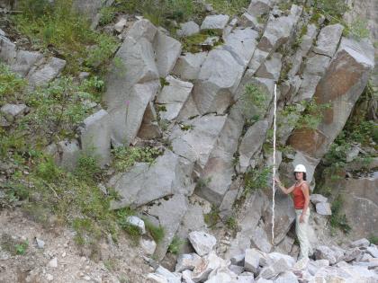 Geklüfteter Granit im Steinbruch der Fa. Fischer-Granit An einem nach links ansteigenden Hang stehen mehrere, nach rechts geneigte und geklüftete Gesteinsreihen. Rechts im Bild, auf losen Steinblöcken stehend, hält eine Frau mit Schutzhelm einen Maßstab an die Gesteinswände.