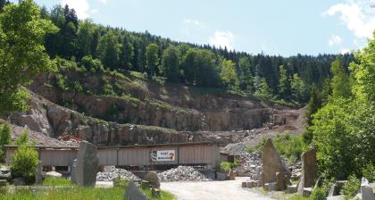 Murgschifferschaftsbruch der Fa. VSG Schwarzwald-Granit-Werke Blick auf ein Steinbruchgelände mit breiten, mittelhohen Gesteinsterrassen und dicht bewaldeter Kuppe. Im Vordergrund Lagergebäude und Gesteinshaufen sowie als Schaustücke aufgestellte Steinblöcke.